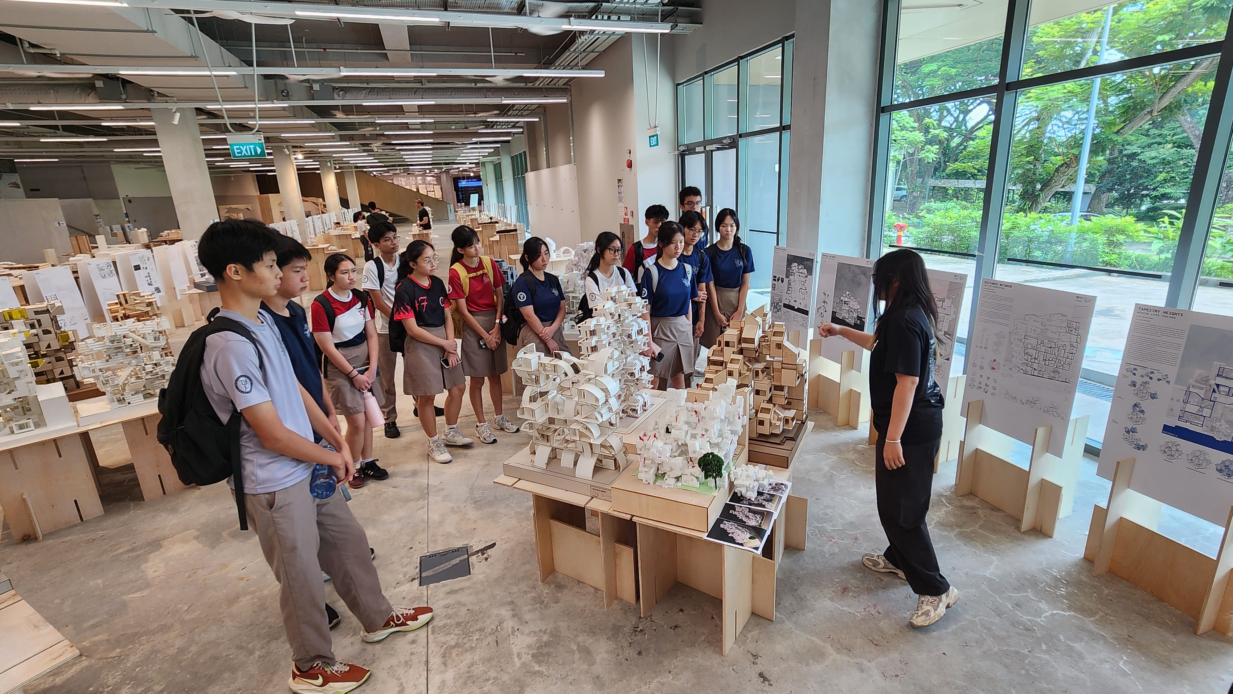 A group of students in school uniforms listens to a guide presenting intricate architectural scale models made of wood and white material, displayed on tables inside a bright, open exhibition space.